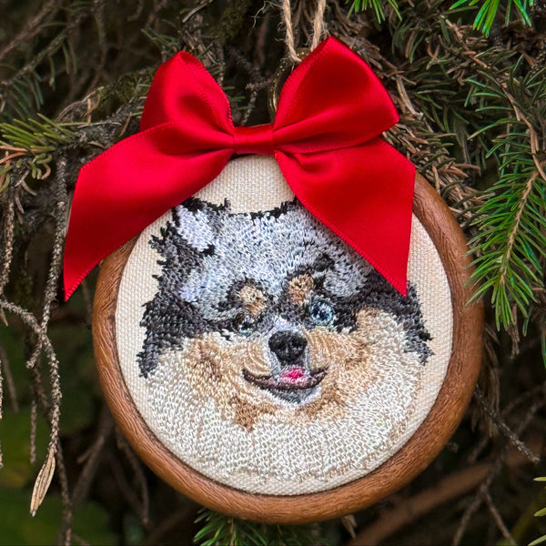 A circular embroidered dog portrait ornament hanging on a Christmas tree. The ornament features a stitched dog face on cream fabric inside a wooden hoop, decorated with a bright red satin bow and hanging twine.