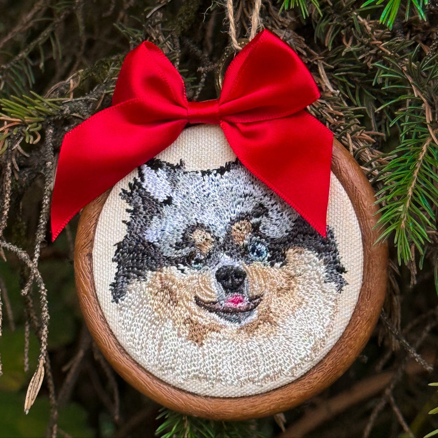 A circular embroidered dog portrait ornament hanging on a Christmas tree. The ornament features a stitched dog face on cream fabric inside a wooden hoop, decorated with a bright red satin bow and hanging twine.