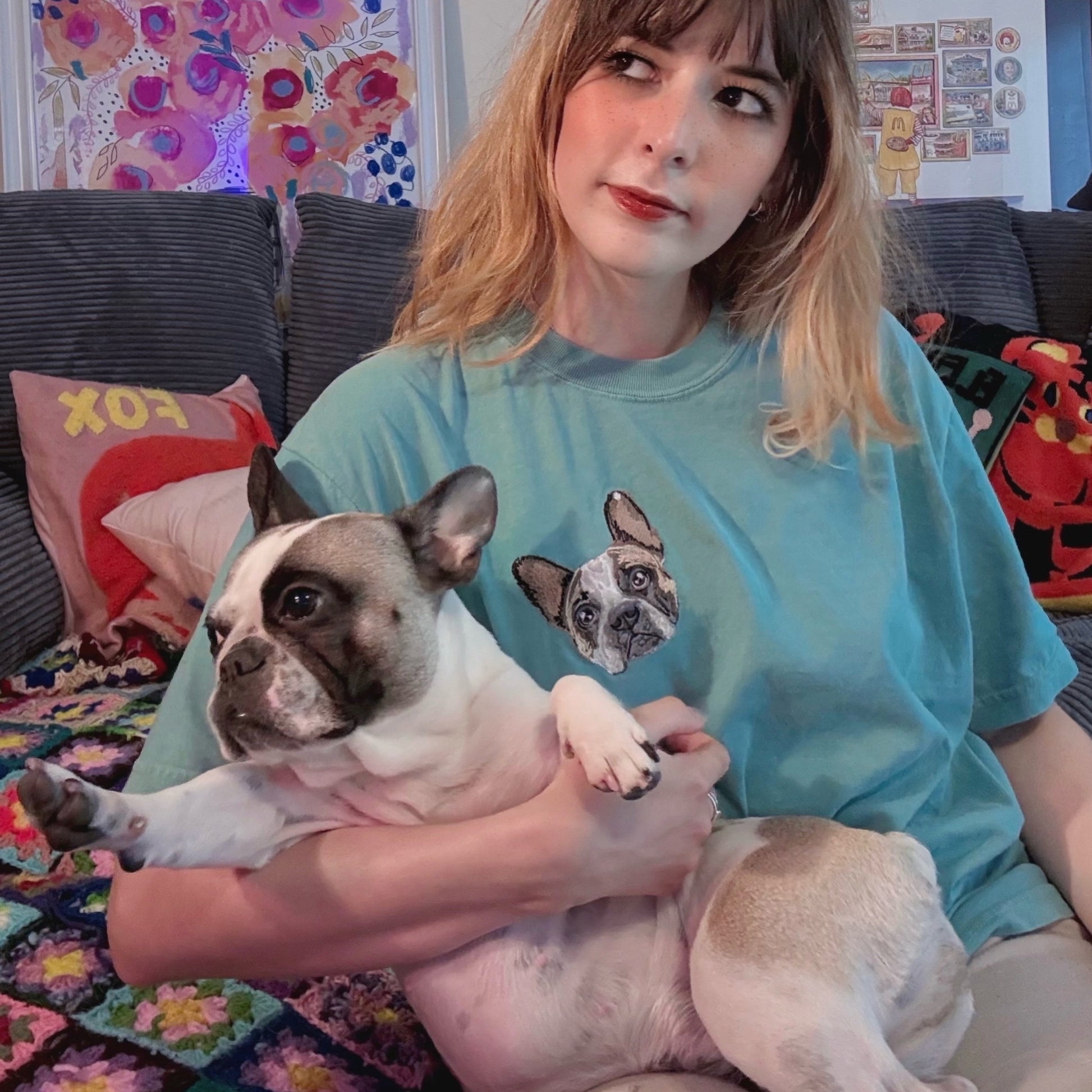 Woman holding a dog wearing a t-shirt with a dog graphic in a colorful living room.