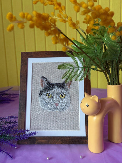 Framed cross-stitched cat portrait on a table with decorative plants and a yellow wall.