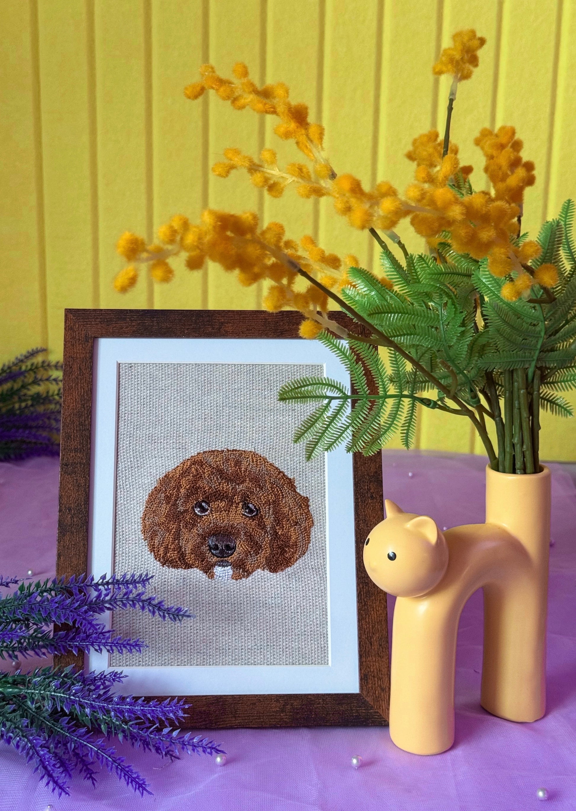 Framed cross-stitched dog portrait on a table with flowers and a vase.