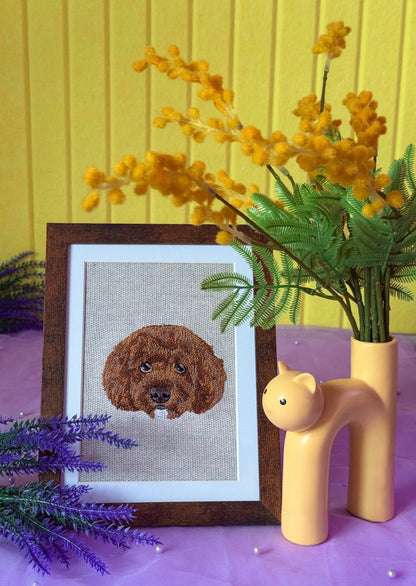 Framed cross-stitched dog portrait on a table with flowers and a vase.