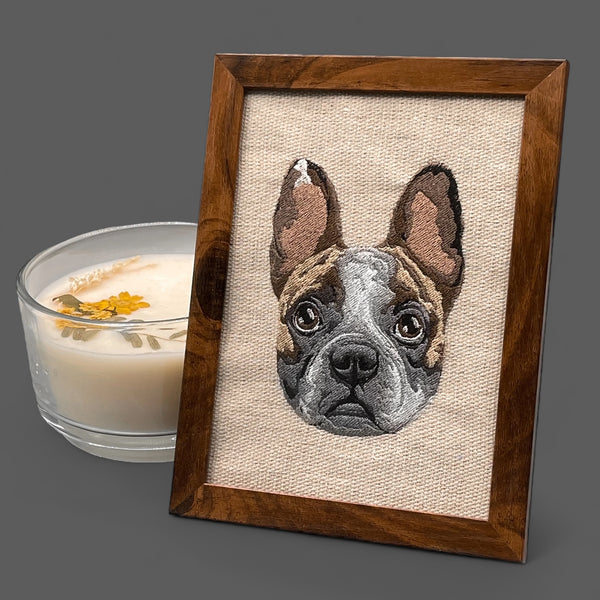 An embroidered canvas picture of a dog's face in a wooden frame, with a candle and a shadow on the side.