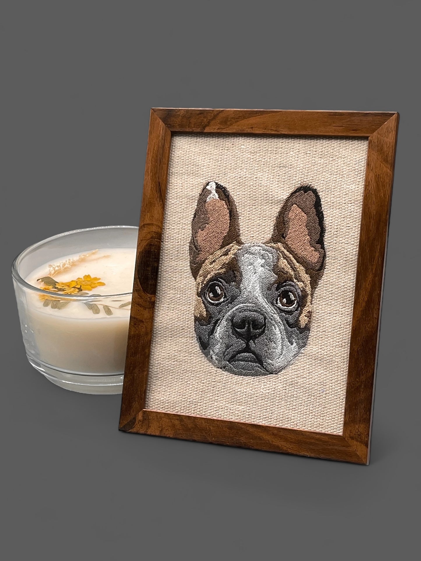 An embroidered canvas picture of a dog's face in a wooden frame, with a candle and a shadow on the side.