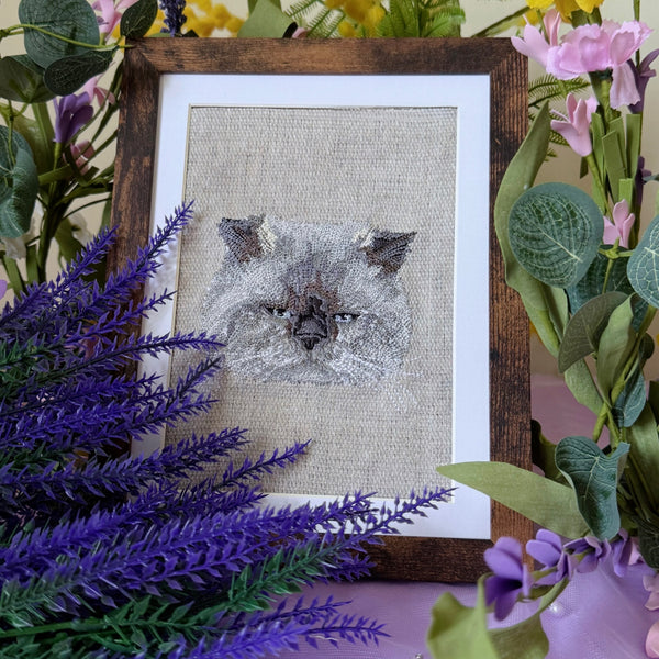 Embroidered cat portrait in a wooden frame surrounded by flowers on a purple surface