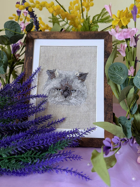 Embroidered cat portrait in a wooden frame surrounded by flowers on a purple surface