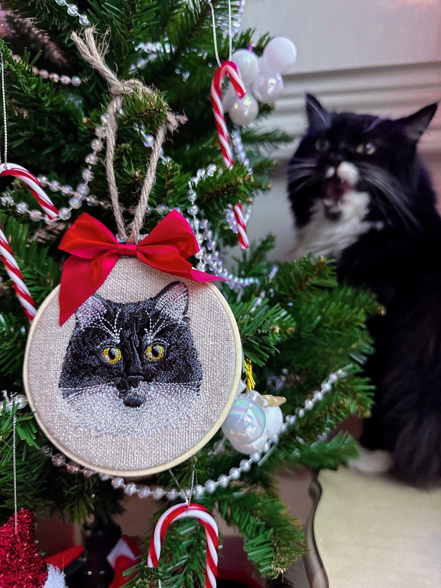 Embroidered cat ornament on a Christmas tree with a cat looking on.