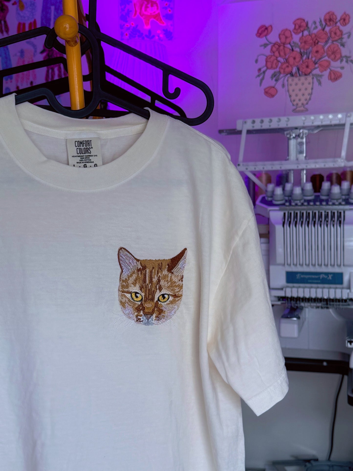White custom pet embroidered t-shirt with a cat graphic on a hanger in a workshop setting.