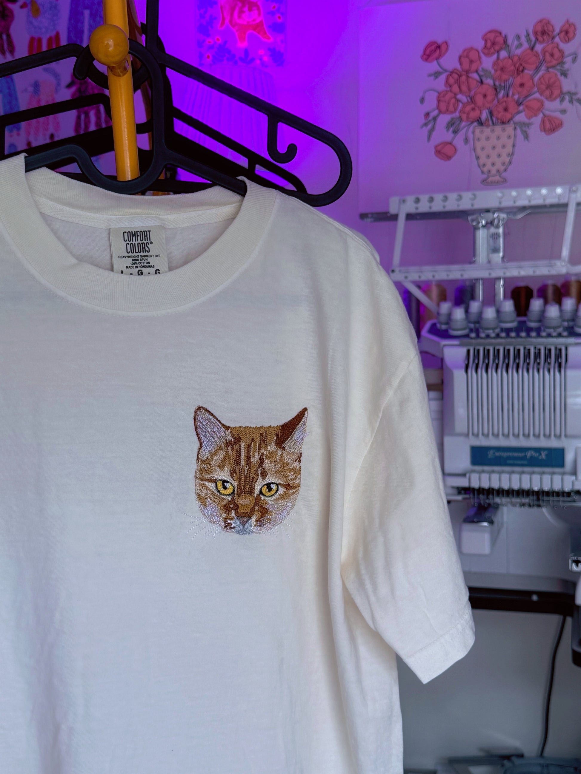 White custom pet embroidered t-shirt with a cat graphic on a hanger in a workshop setting.