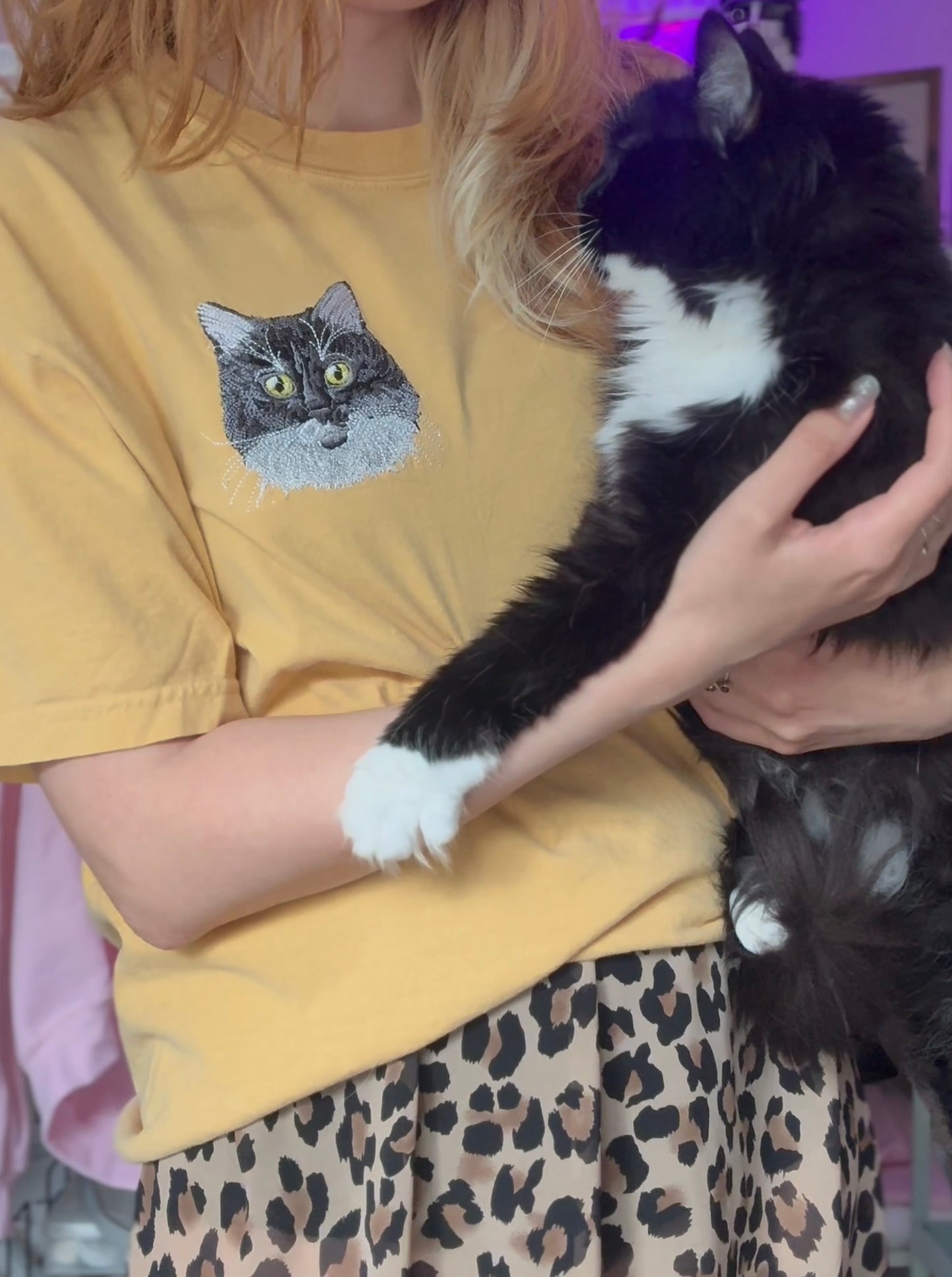 Person wearing a custom pet embroidered yellow shirt with a cat graphic holding a black and white cat.